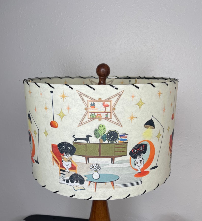 May include: A white lampshade with a black trim and a mid-century modern design featuring black and white dogs, a green plant, a clock, and a bookshelf with a pink flamingo. The lampshade is on a wooden base.