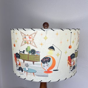 May include: A white lampshade with a black trim features a mid-century modern design with illustrations of dachshunds wearing glasses and reading books in a living room setting. The lampshade has a brown wooden base.
