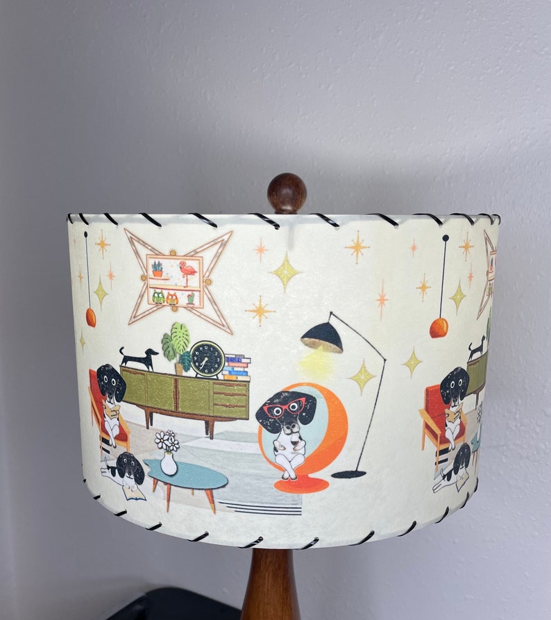 May include: A white lampshade with a black trim features a mid-century modern design with illustrations of dachshunds wearing glasses and reading books in a living room setting. The lampshade has a brown wooden base.