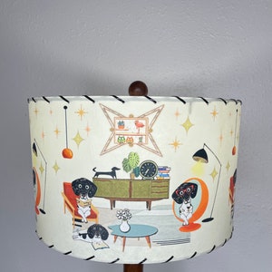 May include: A white lampshade with a black trim features a mid-century modern design with a repeating pattern of dachshunds reading books in a living room setting. The lampshade has a brown wooden base.