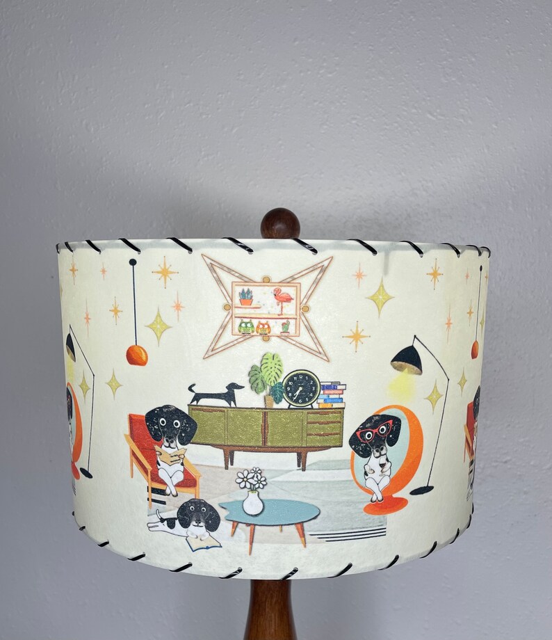 May include: A white lampshade with a black trim features a mid-century modern design with a repeating pattern of dachshunds reading books in a living room setting. The lampshade has a brown wooden base.