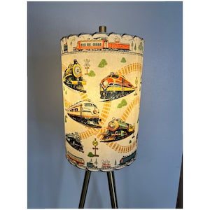 May include: A vintage-style cylindrical lamp with a cream-colored shade featuring colorful illustrations of various trains. The lamp has a tripod base with a brass finish. The train illustrations include yellow, orange, and green locomotives.