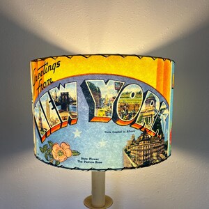 May include: A vintage lampshade featuring a postcard design with the words "Greetings from New York." The shade displays iconic New York landmarks, a floral illustration, and the text "State Flower The Pasture Rose."