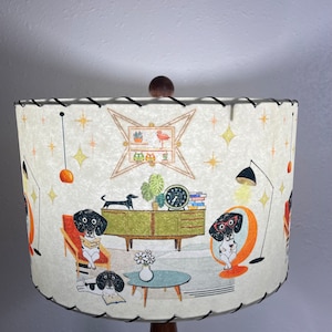 May include: A white lampshade with a mid-century modern design featuring black and white dogs in a living room setting. The lampshade has a brown wooden base and a black trim.