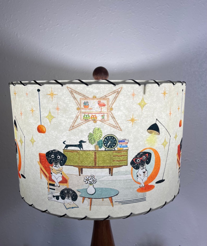 May include: A white lampshade with a mid-century modern design featuring black and white dogs in a living room setting. The lampshade has a brown wooden base and a black trim.