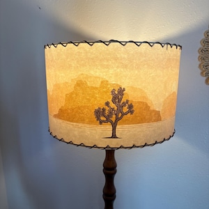 May include: A floor lamp with a cylindrical lampshade featuring a desert landscape design. The shade has a Joshua tree illustration and a stitched black trim. The wooden base is dark brown.