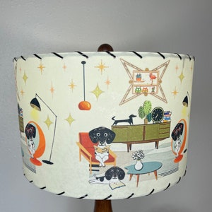 May include: A white lampshade with a mid-century modern design featuring black and white dogs reading books in a living room setting. The lampshade has a black trim and a brown wooden base.