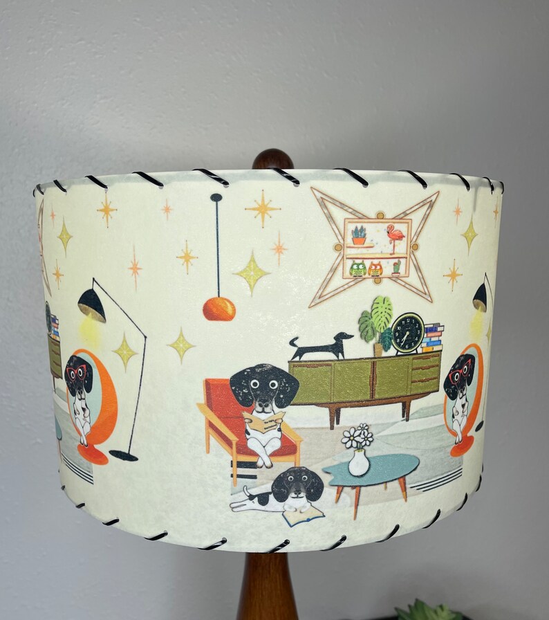 May include: A white lampshade with a mid-century modern design featuring black and white dogs reading books in a living room setting. The lampshade has a black trim and a brown wooden base.