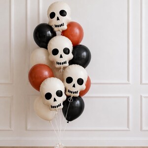 May include: A balloon bouquet featuring three off-white skull-shaped balloons with black facial features, interspersed with round balloons in solid black, burnt orange, and cream colors, all tied together with white ribbons at the base.
