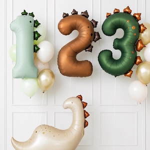 May include: A set of foil balloons shaped like the numbers 1, 2, and 3, decorated with dark green, brown, and copper dinosaur spikes. A beige dinosaur-shaped foil balloon stands below, accompanied by round white and gold latex balloons.
