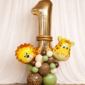 Könnte beinhalten: Eine goldene Folienballon-Nummer eins mit Löwen- und Giraffenballons. Das Arrangement umfasst grüne, braune und weiße Ballons und schafft ein Safari- oder Dschungel-Thema für eine erste Geburtstagsfeier.