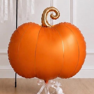 May include: A large, round, orange pumpkin-shaped foil balloon with a metallic gold curly stem. The balloon is anchored at the bottom by a white ribbon tied in a bow, which is attached to a small, round, white weighted object on the floor.