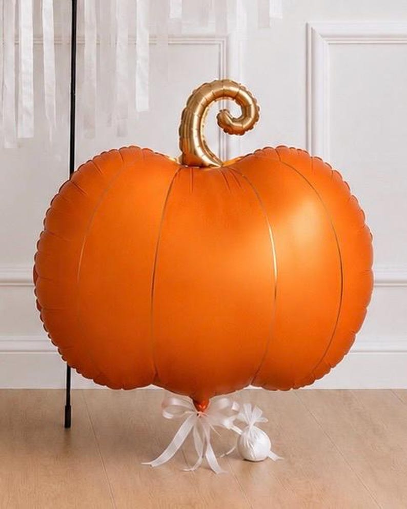 May include: A large, round, orange pumpkin-shaped foil balloon with a metallic gold curly stem. The balloon is anchored at the bottom by a white ribbon tied in a bow, which is attached to a small, round, white weighted object on the floor.