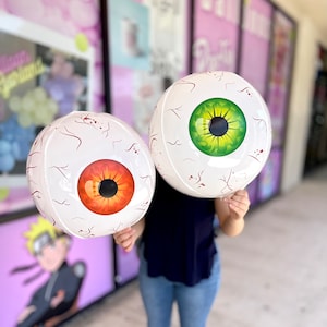 Puede incluir: Dos globos inflables de ojos blancos, uno con un iris verde y el otro con un iris naranja. Los globos están siendo sostenidos por una persona.