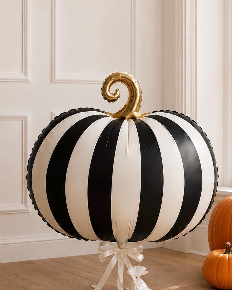 May include: A large, round, inflatable pumpkin balloon featuring vertical black and white stripes. The pumpkin has a metallic gold, spiraled stem at the top and is tied at the base with a white ribbon. The surface has a smooth, glossy finish.