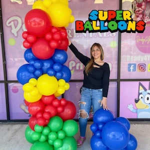 Könnte beinhalten: Eine hohe Säule aus roten, gelben, blauen und grünen Luftballons. Die Ballons sind in einem gestapelten Muster angeordnet und bilden ein farbenfrohes und festliches Display. Der Text "SUPER BALLOONS" ist im Hintergrund sichtbar.