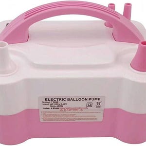 Könnte beinhalten: Eine weiß-rosa elektrische Ballonpumpe mit einem Tragegriff, zwei Aufblasdüsen und einem Etikett mit der Aufschrift ELECTRIC BALLOON PUMP, Model: 7 2085, Input: AC 220V-240V, Watt: 600W, Noise: < 85dB, indoor and household use only, CAUTION.