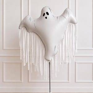 May include: A white ghost-shaped foil balloon featuring a black printed face with two oval eyes and an oval mouth. The balloon is adorned with long, white ribbon streamers hanging from its base and is attached to a thin black pole with a white base.