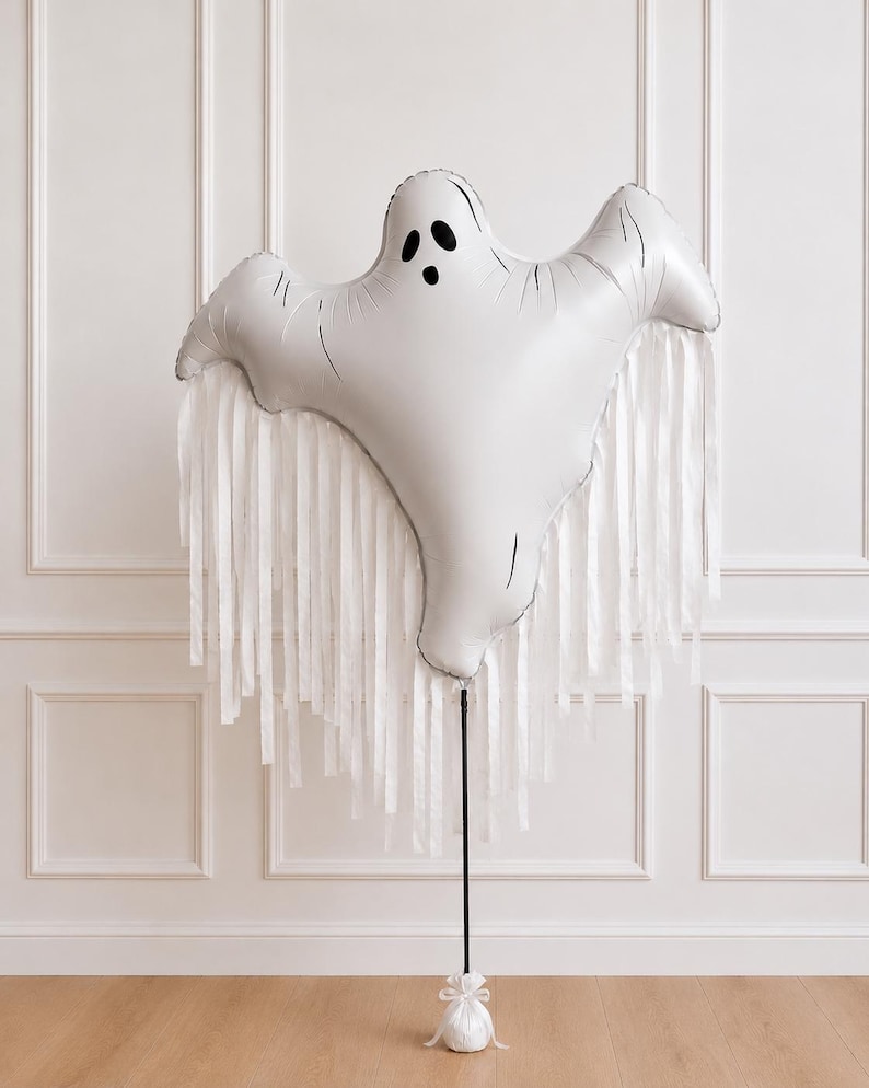 May include: A white ghost-shaped foil balloon featuring a black printed face with two oval eyes and an oval mouth. The balloon is adorned with long, white ribbon streamers hanging from its base and is attached to a thin black pole with a white base.