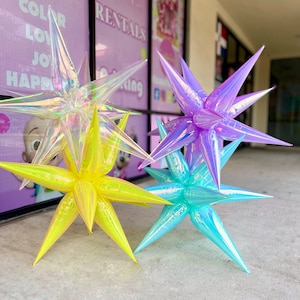 Puede incluir: Tres globos iridiscentes en forma de estrella en amarillo, morado y azul. Los globos están inflados y tienen una punta puntiaguda en cada punto de la estrella.