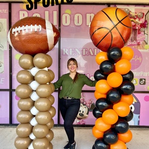 Puede incluir: Dos columnas de globos decoradas con globos de temática deportiva. La columna de la izquierda tiene globos marrones y blancos con un balón de fútbol en la parte superior. La columna de la derecha tiene globos naranjas y negros con un balón de baloncesto en la parte superior. El texto "SPORTS COLUMNS" se muestra encima de las columnas.