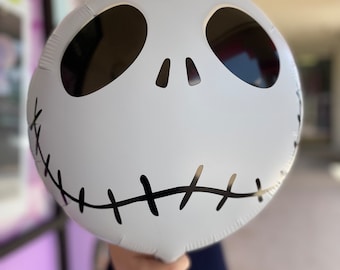 Puede incluir: Un globo blanco con forma de la cara de Jack Skellington de Pesadilla antes de Navidad. El globo tiene ojos negros, una nariz negra y una boca negra cosida.