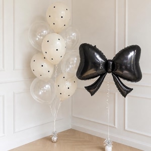 May include: A cluster of balloons featuring cream-colored balloons with black polka dots and clear balloons, anchored by a silver weight, alongside a large, singular black foil balloon shaped like a bow, also anchored by a silver weight.