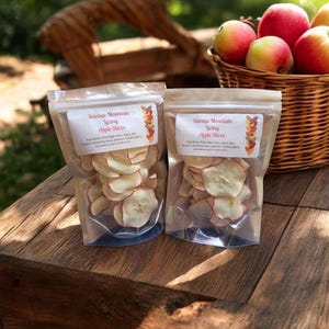 May include: Two clear plastic bags of apple slices with red edges sit on a rustic wooden table. Each bag has a label that reads "Savage Mountain Living Apple Slices." A wicker basket filled with red and yellow apples is in the background.