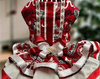 May include: A red and white pet dress featuring University of Alabama Crimson Tide logos, football graphics, and the text Alabama. The dress includes a layered skirt with silver ribbon trim and a central white bow with pearl-like accents.