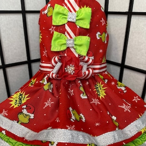Puede incluir: Vestido rojo y verde para perro con estampado de Grinch, cinta a rayas blancas y rojas y lazo verde. El vestido tiene un ribete de purpurina plateada.