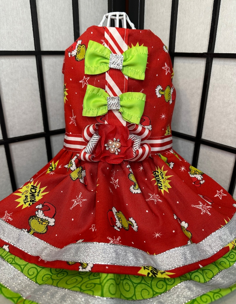 Puede incluir: Vestido rojo y verde para perro con estampado de Grinch, cinta a rayas blancas y rojas y lazo verde. El vestido tiene un ribete de purpurina plateada.
