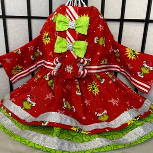 Puede incluir: Vestido de Navidad rojo y verde para una mascota con estampado de Grinch, ribete a rayas blanco y rojo y un lazo verde. El vestido tiene una capa inferior verde y blanca con ribete plateado.