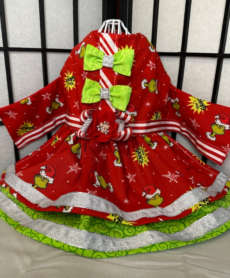 Puede incluir: Vestido de Navidad rojo y verde para una mascota con estampado de Grinch, ribete a rayas blanco y rojo y un lazo verde. El vestido tiene una capa inferior verde y blanca con ribete plateado.