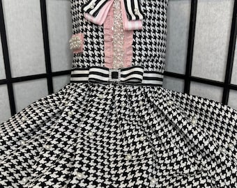 May include: A black and white houndstooth dog dress with a pink and white bow, pearl accents, and a black and white striped ribbon.
