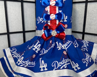 May include: A blue and white dog dress with a red and white ribbon. The dress has the Los Angeles Dodgers logo and baseballs printed on it.