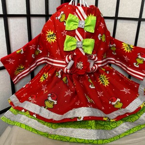 Puede incluir: Vestido rojo y verde para perro con estampado de Grinch. El vestido tiene una cinta blanca y roja a rayas alrededor de la cintura y dos lazos verdes en el corpi&ntilde;o. El vestido tiene una falda de capas con ribete plateado.