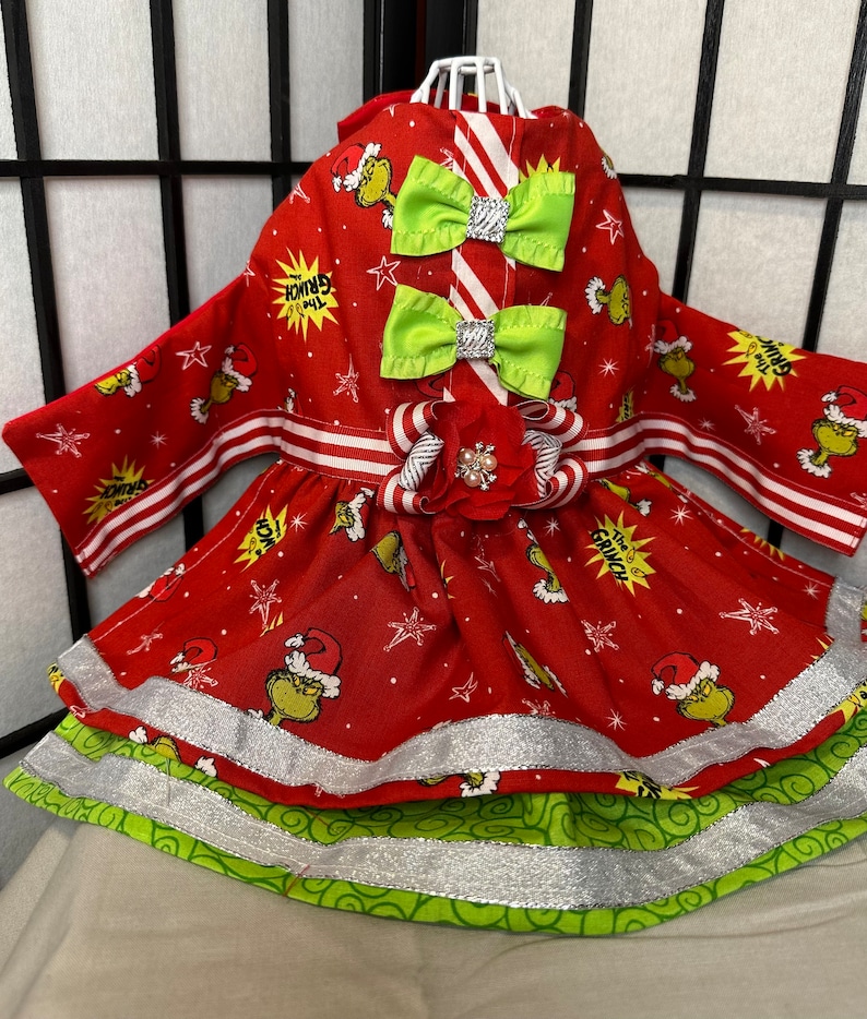 Puede incluir: Vestido rojo y verde para perro con estampado de Grinch. El vestido tiene una cinta blanca y roja a rayas alrededor de la cintura y dos lazos verdes en el corpi&ntilde;o. El vestido tiene una falda de capas con ribete plateado.