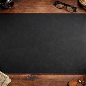 May include: A black rectangular desk mat with red stitching, surrounded by office supplies on a wooden desk. Items include a camera, glasses, a pen, a cup of coffee, and a wallet.