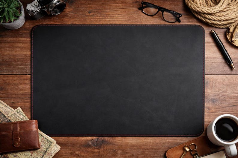 May include: A black rectangular desk mat with red stitching, surrounded by office supplies on a wooden desk. Items include a camera, glasses, a pen, a cup of coffee, and a wallet.