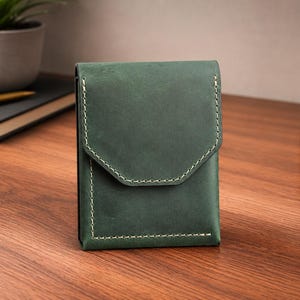 May include: A dark green leather wallet with a flap closure and white stitching. The wallet is rectangular and sits on a wooden surface. A notebook, pen, and plant are in the background.