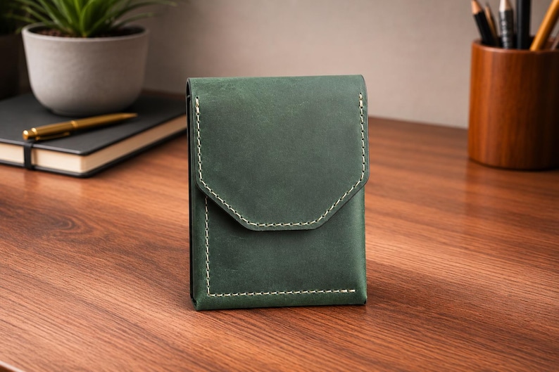 May include: A dark green leather wallet with a flap closure and white stitching. The wallet is rectangular and sits on a wooden surface. A notebook, pen, and plant are in the background.
