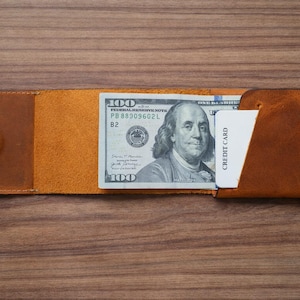 May include: A handmade, open leather wallet in shades of brown, holding a one-hundred-dollar bill and a white credit card. The wallet is open, displaying the bill and card, and is set against a wooden surface.