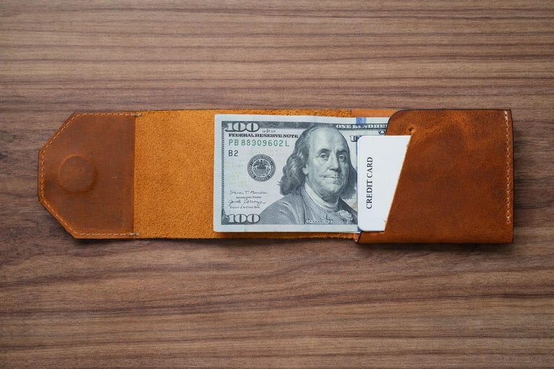 May include: A handmade, open leather wallet in shades of brown, holding a one-hundred-dollar bill and a white credit card. The wallet is open, displaying the bill and card, and is set against a wooden surface.