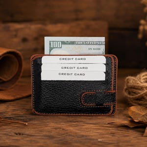 May include: A black textured leather card holder with brown stitched edges, featuring three horizontal slots containing white cards labeled CREDIT CARD and a central compartment holding a one hundred dollar bill with visible text.