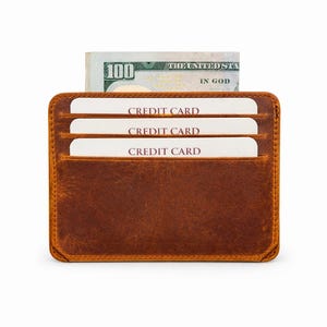 May include: A brown leather card holder featuring three horizontal slots containing white cards labeled CREDIT CARD and a central compartment holding a one hundred dollar bill with the text THE UNITED STATES OF AMERICA IN GOD.