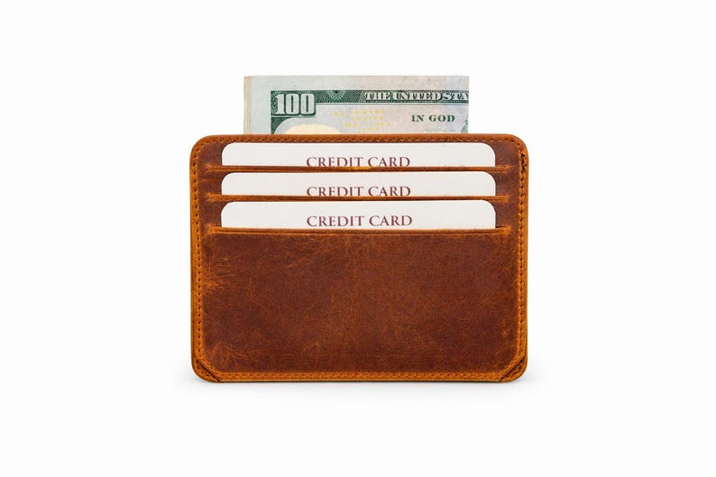 May include: A brown leather card holder featuring three horizontal slots containing white cards labeled CREDIT CARD and a central compartment holding a one hundred dollar bill with the text THE UNITED STATES OF AMERICA IN GOD.