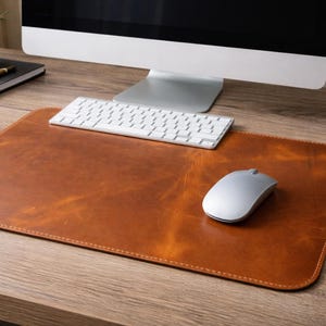 May include: A brown leather desk mat with a stitched border, a silver computer mouse, and a white keyboard. The desk mat is rectangular and sits on a wooden desk. A computer monitor is in the background.