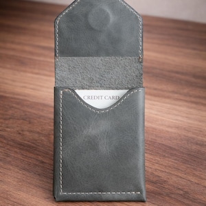 May include: A gray leather card holder with white stitching and a magnetic closure. The interior reveals a white card with the words "CREDIT CARD". The holder is open, displaying the card. The background is a wooden surface.