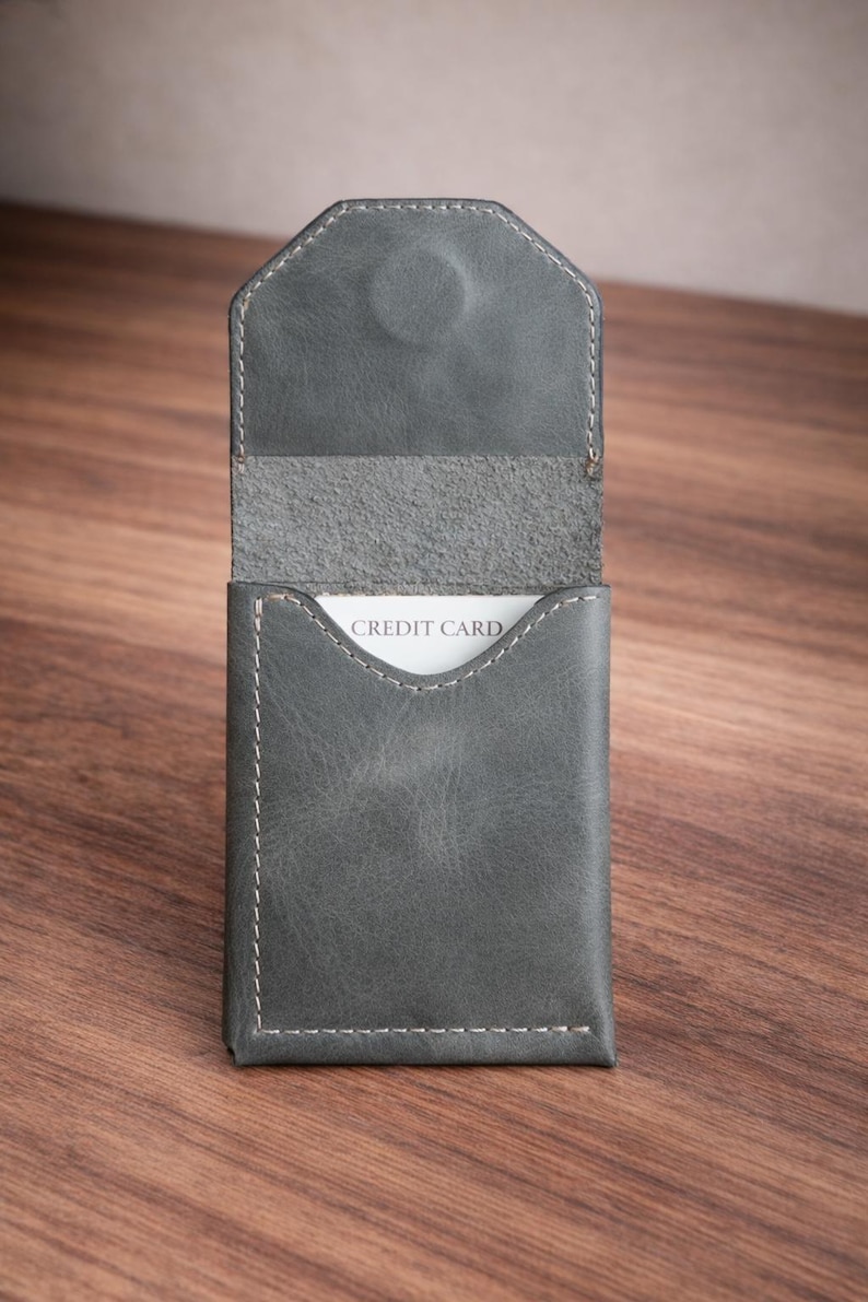 May include: A gray leather card holder with white stitching and a magnetic closure. The interior reveals a white card with the words "CREDIT CARD". The holder is open, displaying the card. The background is a wooden surface.