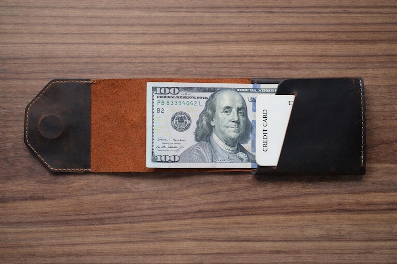 May include: A handmade leather wallet in shades of brown and black, open to display a one hundred dollar bill and a credit card. The wallet is on a wooden surface.